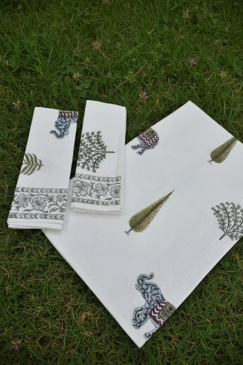 A set of white cotton towels featuring hand-block printed elephant and leaf motifs in muted green, gray, and purple, laid out on a green grass background.