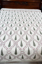 A white dohar featuring a repeating green and red geometric and floral pattern, laid out on a bed with a dark wooden headboard visible at the top.