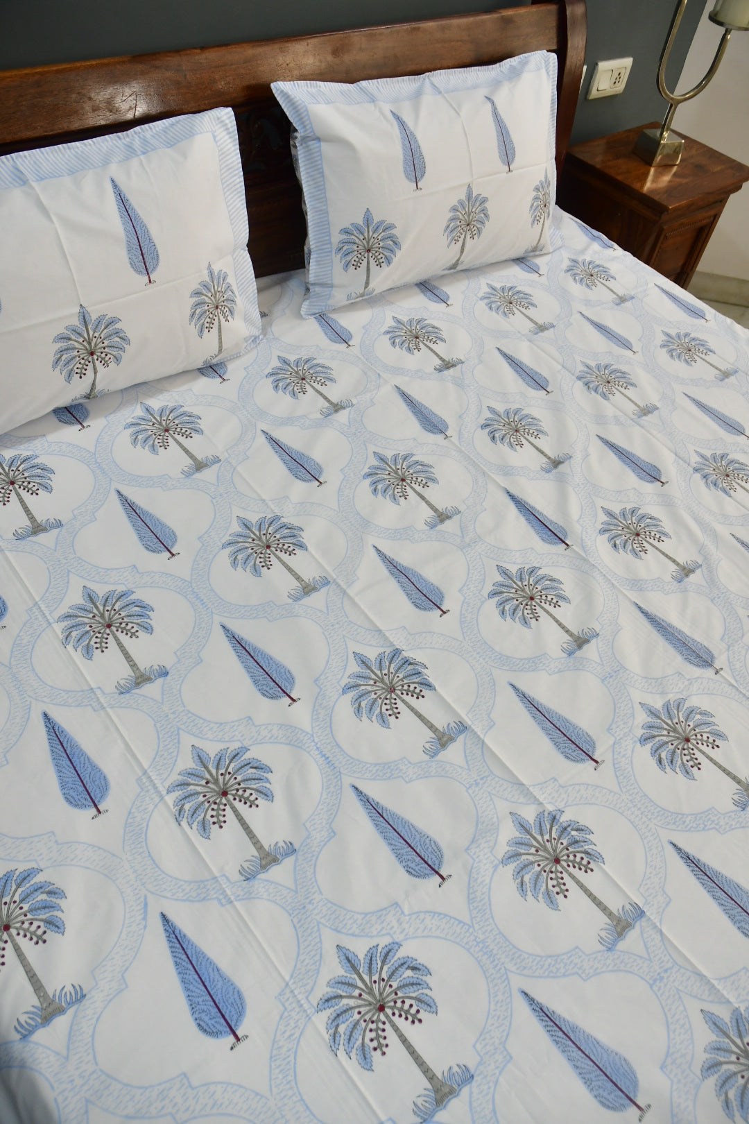 A bed dressed with a white bedsheet  featuring a repeating light blue palm tree and teardrop pattern, with two matching pillowcases, shown in a bedroom setting with a wooden headboard and bedside table.