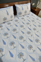 A bed dressed with a white bedsheet  featuring a repeating light blue palm tree and teardrop pattern, with two matching pillowcases, shown in a bedroom setting with a wooden headboard and bedside table.