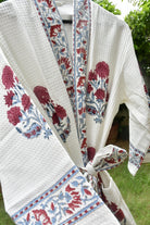 A white textured cotton kimono robe with red and blue floral embroidery, tied at the waist, shown outdoors with green foliage in the background.