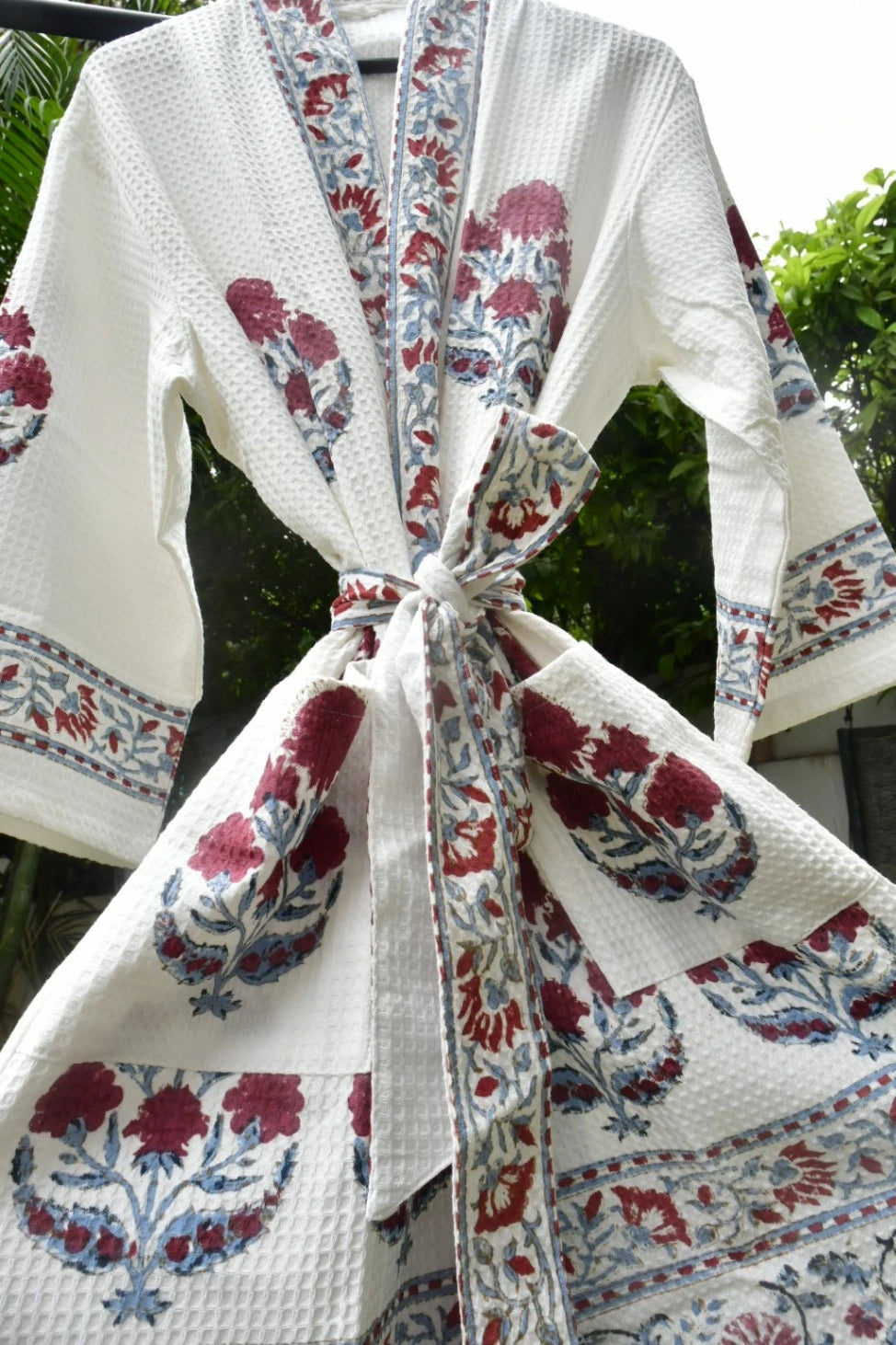 A white waffle-weave cotton robe with red and blue floral block prints and a matching tie belt, shown hanging outdoors against a background of green foliage.
