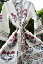 A white waffle-weave cotton robe with red and blue floral block prints and a matching tie belt, shown hanging outdoors against a background of green foliage.