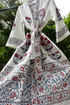 A white cotton block-print robe with red and blue floral motifs, hanging outdoors against a background of green foliage.