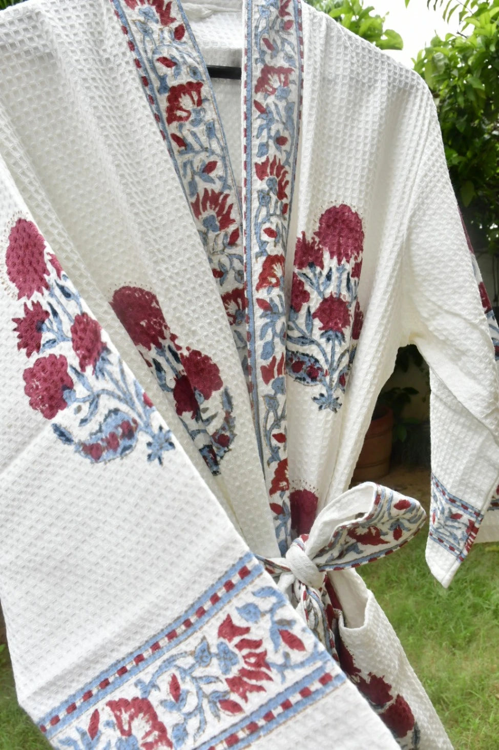A white textured cotton robe with red and blue floral block prints and border patterns, tied at the waist, shown outdoors with green foliage in the background.