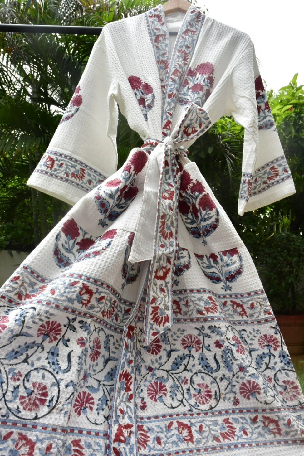 A white cotton kimono robe with red and blue floral block prints, tied at the waist, hanging outdoors against a background of green foliage.
