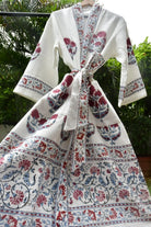 A white cotton kimono robe with red and blue floral block prints, tied at the waist, hanging outdoors against a background of green foliage.