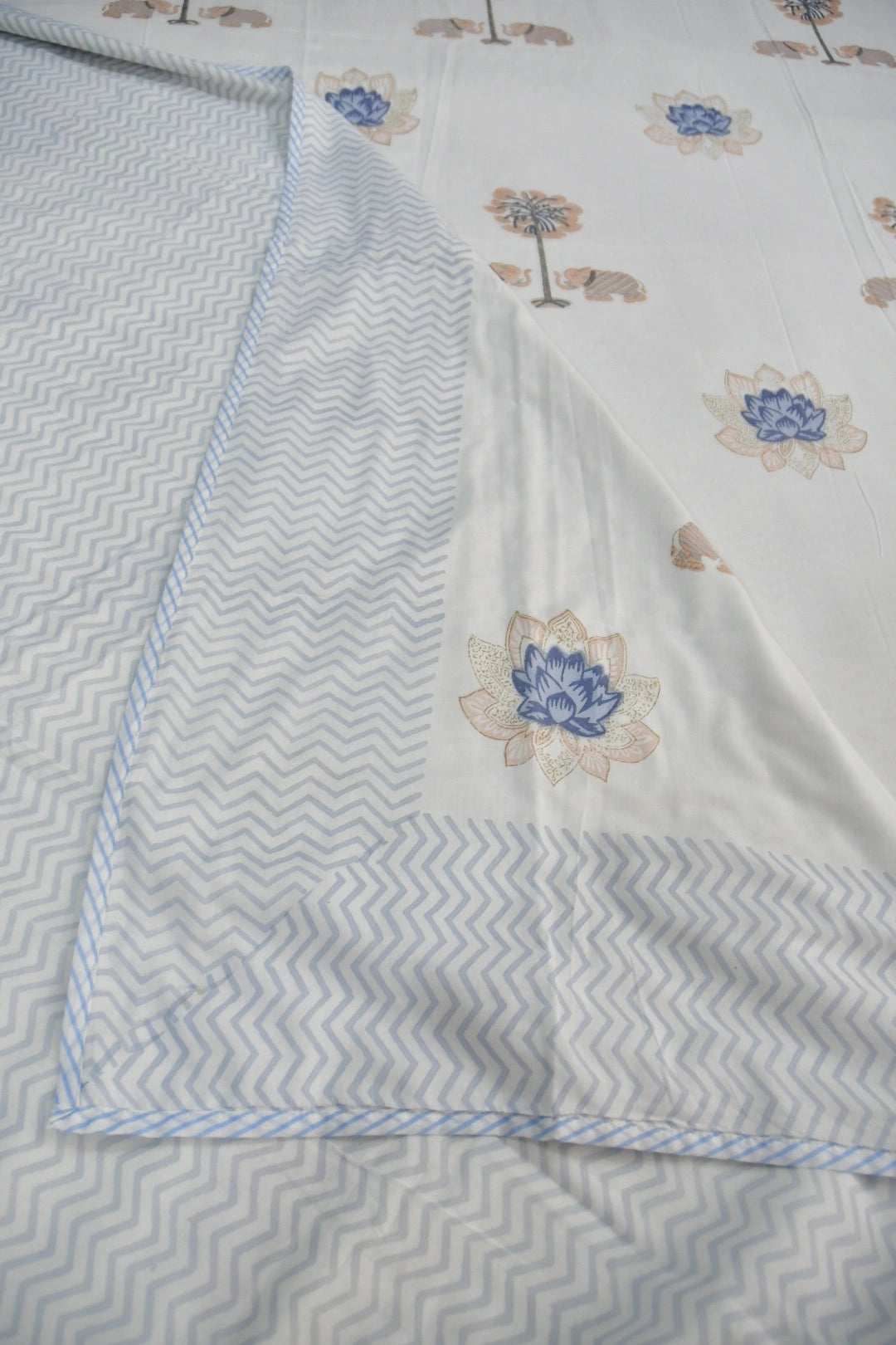 A folded white fabric item, likely a bed sheet or duvet cover, featuring a light blue and white chevron pattern on one side and a white base with blue and beige floral motifs on the other.