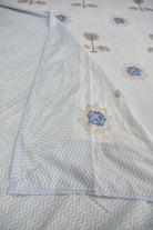 A folded white fabric item, likely a bed sheet or duvet cover, featuring a light blue and white chevron pattern on one side and a white base with blue and beige floral motifs on the other.