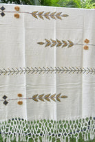 An off-white cotton throw blanket with bohemian-style embroidery and a fringed hem, draped outdoors over a white frame with green foliage visible in the background.