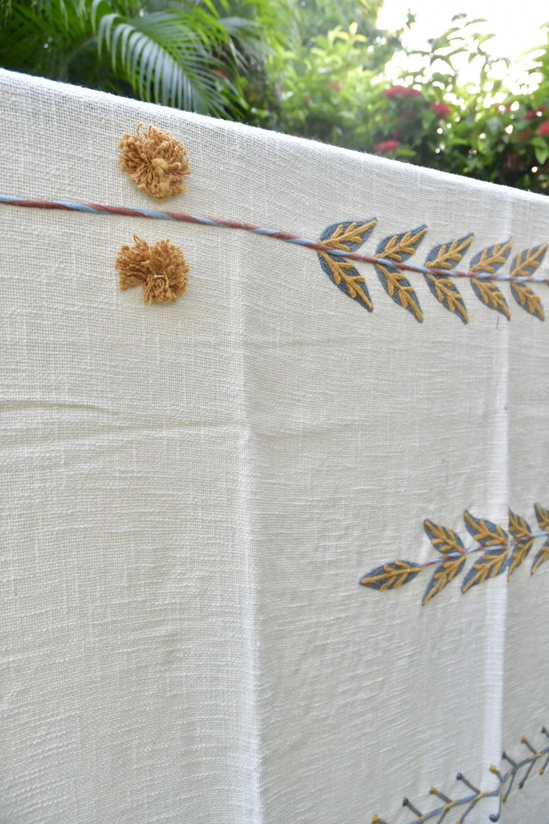An off-white linen tablecloth featuring embroidered leaf patterns in gold and blue, with two decorative gold tassels, shown outdoors with green foliage in the background.