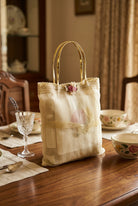 A square beige fabric pillow with a gold metal handle and a subtle embroidered or printed floral motif in the center, resting on a dark surface.