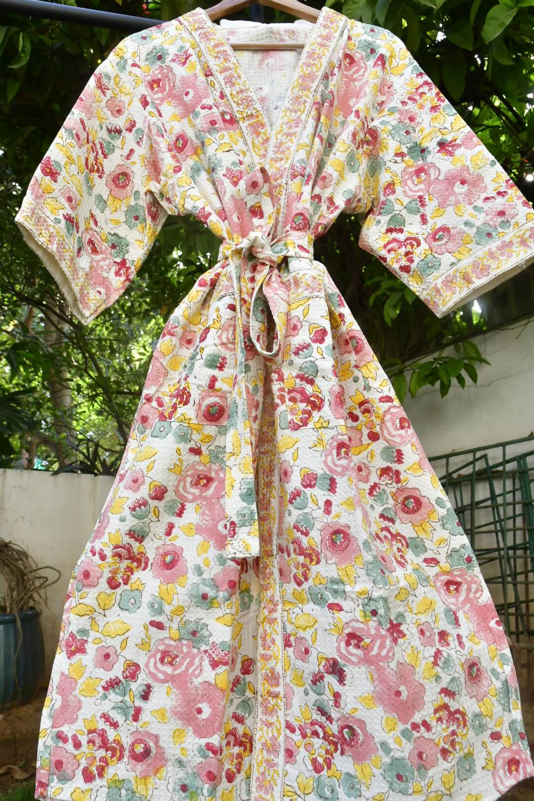 A floral print wrap robe with a tie waist, hanging on a wooden hanger outdoors against a background of green foliage and a white wall.