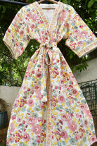 A floral print wrap robe with a tie waist, hanging on a wooden hanger outdoors against a background of green foliage and a white wall.