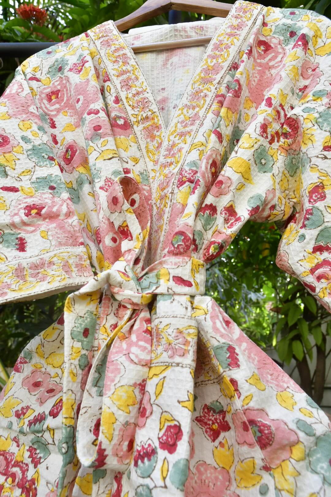 A close-up of a floral kimono-style robe or wrap hanging on a hanger outdoors. The fabric has a cream base with pink, red, yellow, and green floral print, and the robe is tied at the waist.