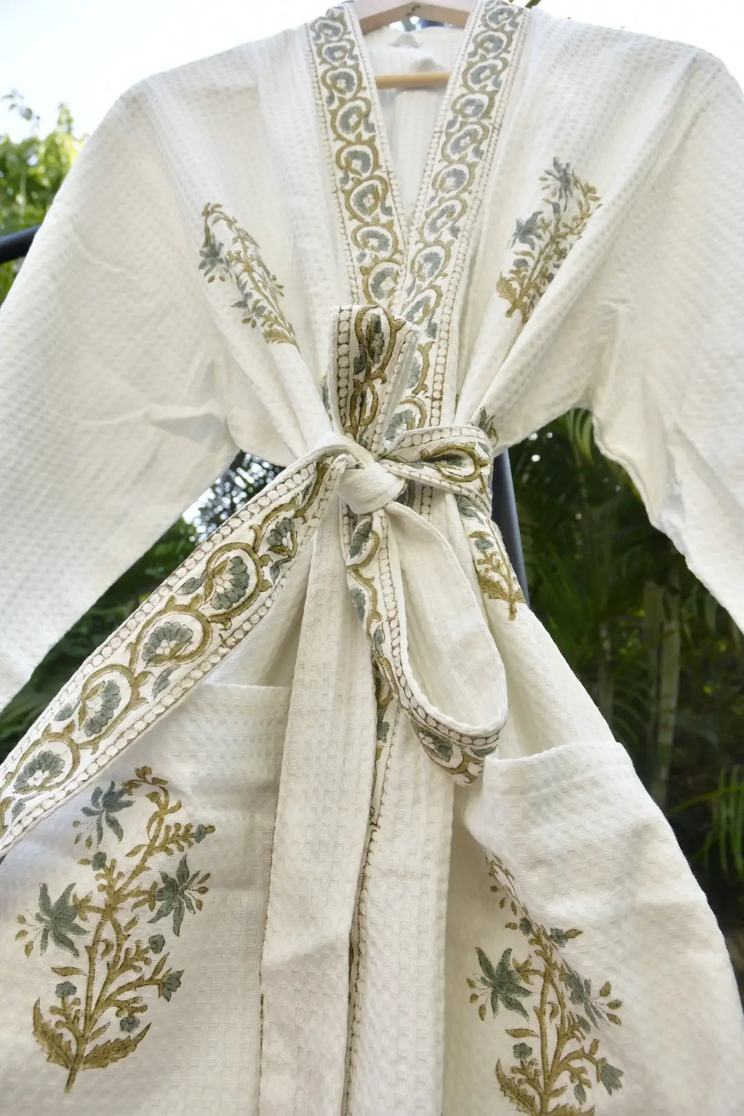 White embroidered garment with gold and green details on a hanger against a blurred natural background.