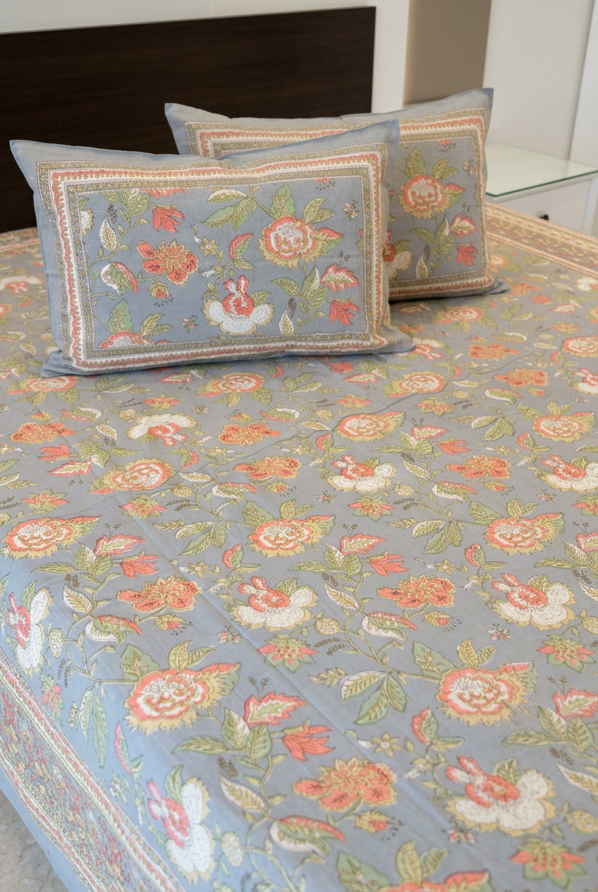 A floral-patterned bedspread or duvet set displayed on a bed, with matching decorative pillows. The fabric features a dusty blue-gray base color with a dense print of pink, orange, green, and white flowers.