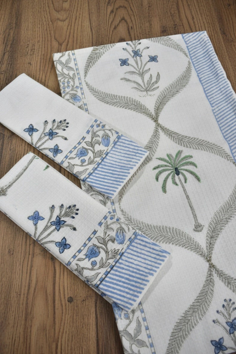 A set of white cotton napkins with blue and green block-printed floral and palm tree motifs, arranged on a wooden tabletop.