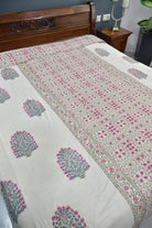A cream-colored dohar / AC blanket with pink and green block-print floral motifs and borders, shown on a bed with a dark wood headboard in a bedroom setting.