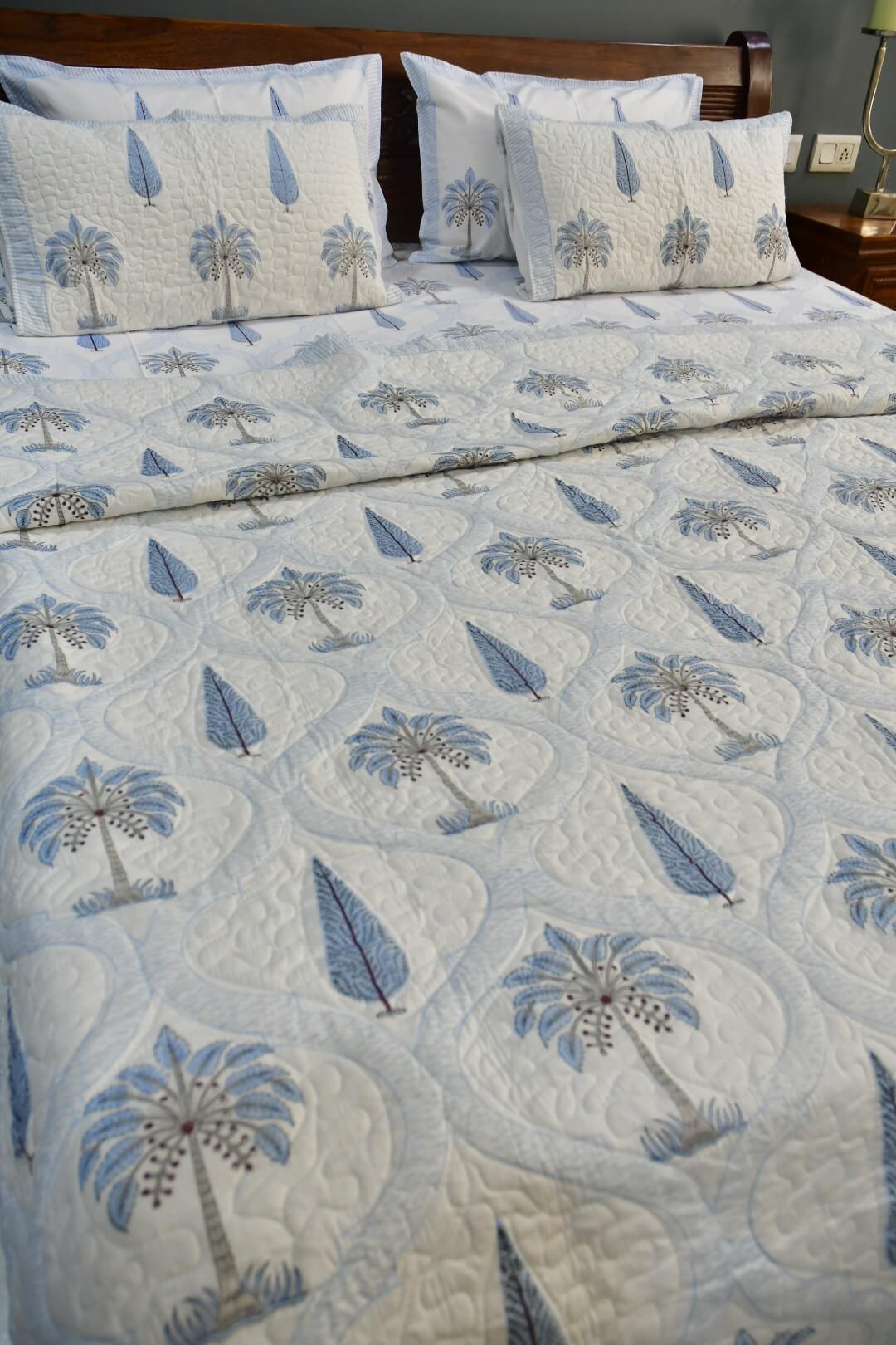 A quilted bedspread and matching pillow shams featuring a blue and gray palm tree and leaf pattern on a white background, styled on a bed with a wooden headboard.