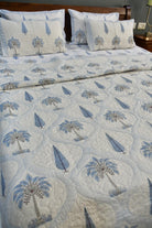 A quilted bedspread and matching pillow shams featuring a blue and gray palm tree and leaf pattern on a white background, styled on a bed with a wooden headboard.