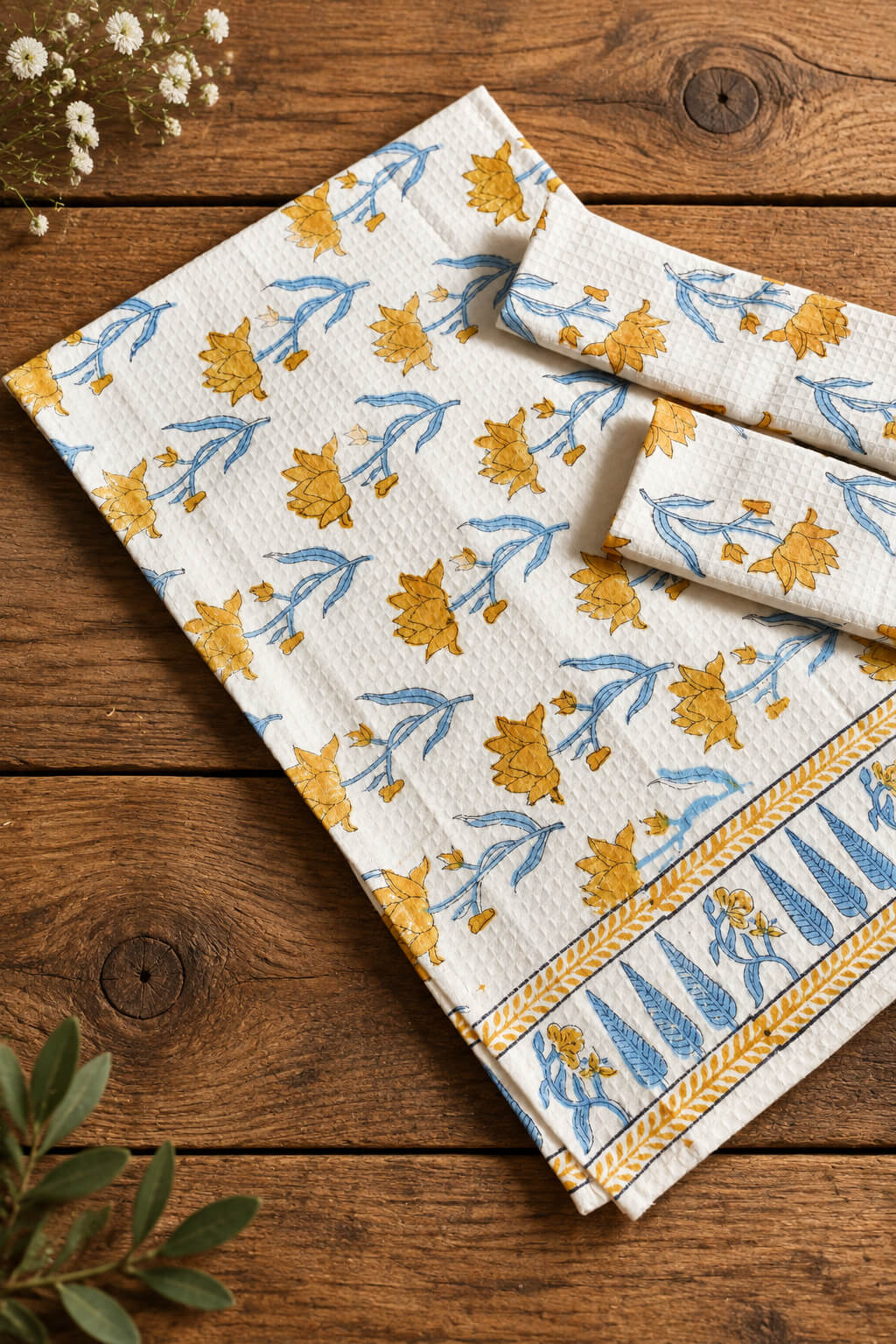 A folded white cotton tea towel with a yellow and blue floral block print pattern, displayed on a rustic wooden surface alongside a matching folded napkin.