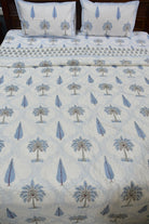 A bed made with a white quilt or duvet featuring a repeating blue palm tree and teardrop pattern, with matching pillowcases and a patterned border visible along the top.