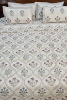 An off-white quilted bedspread with a repeating floral and geometric trellis pattern in beige, blue, and purple, styled on a bed with matching embroidered pillows.