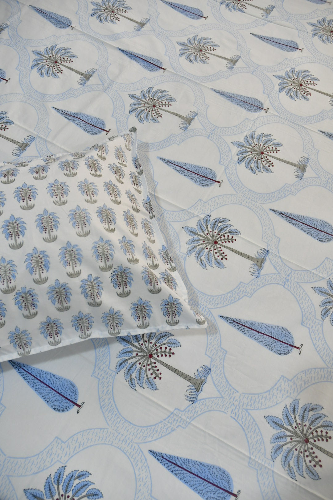 A close-up of a white bedsheet featuring a printed sheet and matching pillowcase with a blue and gray palm tree and leaf pattern.