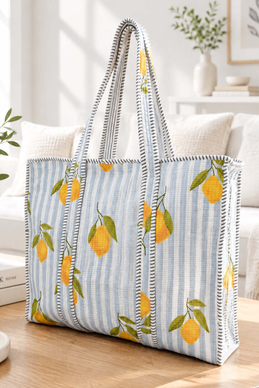 A tote bag with a light blue and white striped fabric body, decorated with yellow lemons and green leaves, featuring black and white striped handles, set on a wooden table in a bright living room.