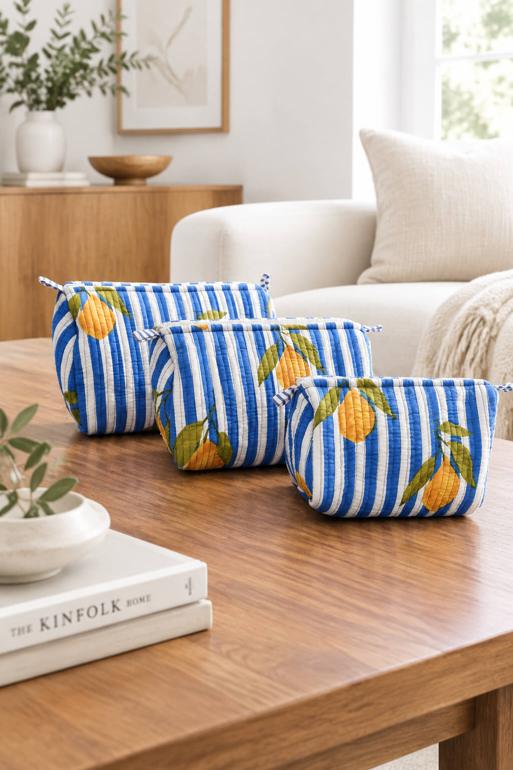 Three zippered cosmetic pouches with a blue and white striped pattern and orange lemon print, arranged on a wooden table in a bright living room setting.