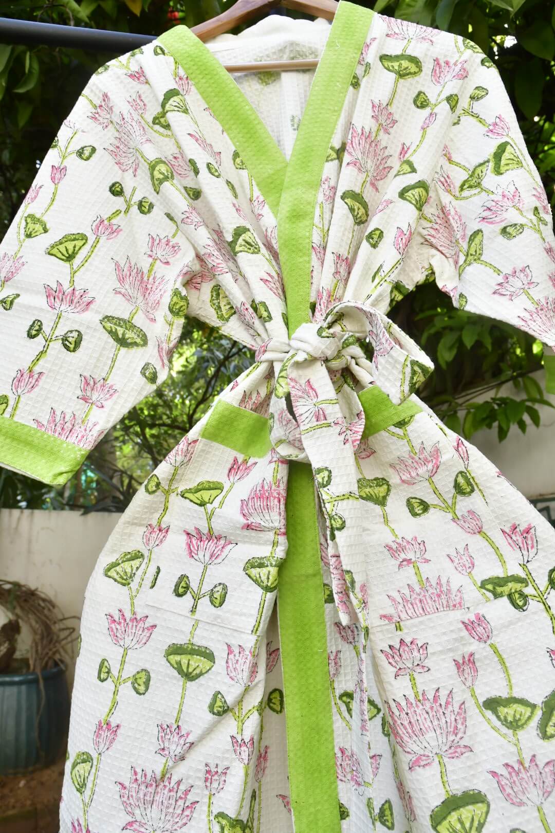 A white cotton robe with a pink lotus and green lotus leaf pattern, featuring lime green trim and a matching tie belt, hanging on a wooden hanger outdoors.