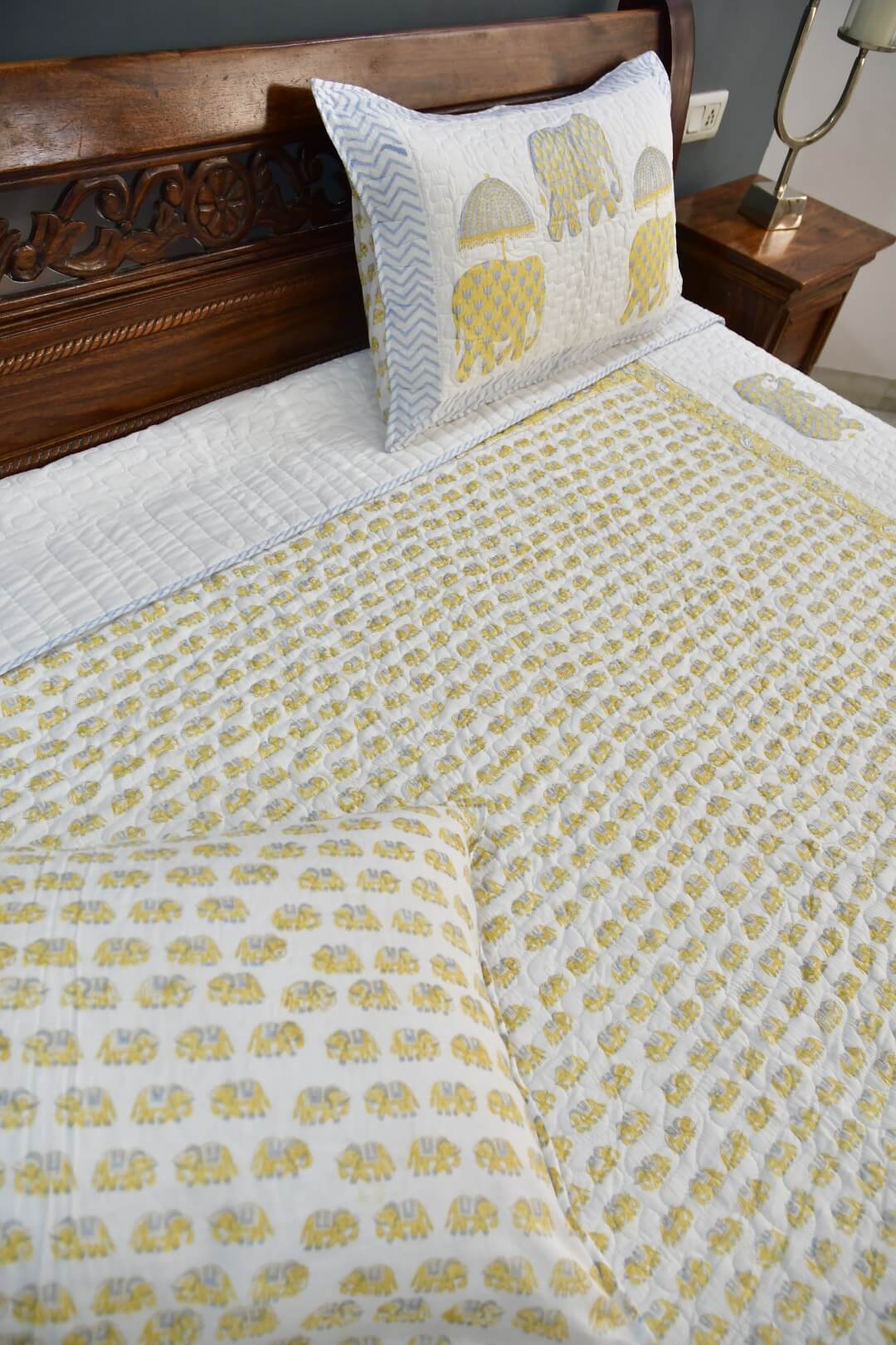 A quilted bedding set featuring a white quilt with a yellow elephant pattern, a matching pillowcase with chevron stripes and yellow elephants, and a dark wood headboard in a bedroom setting.