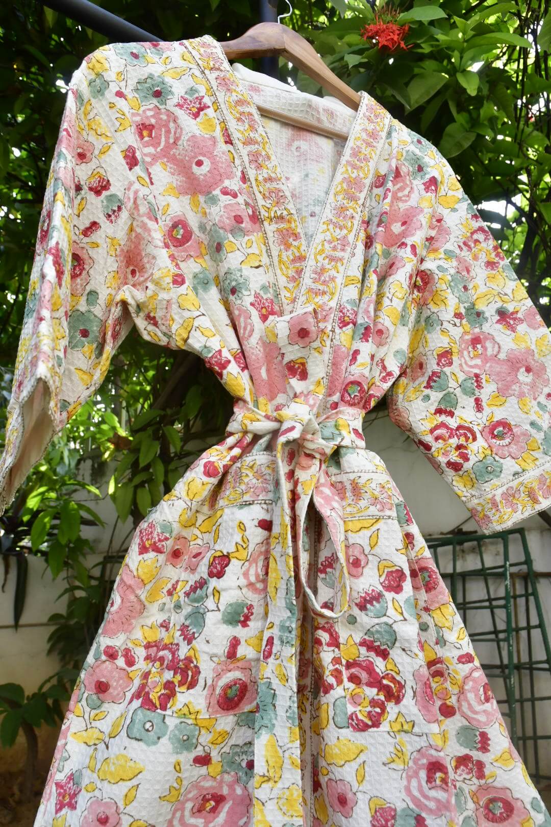 A floral print kimono-style robe with a tie waist, hanging on a wooden hanger outdoors against a background of green foliage.