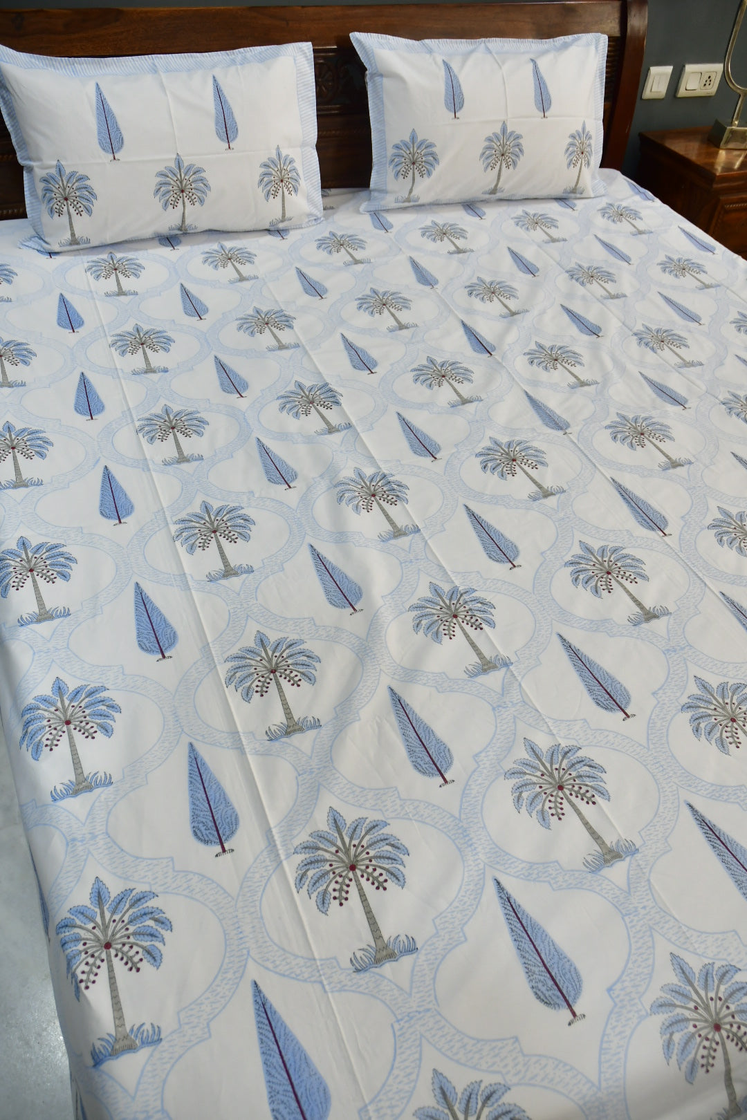 A bed dressed with a white sheet or duvet featuring a repeating blue and gray palm tree and leaf pattern, with two matching pillowcases against a dark wooden headboard.