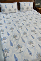 A bed dressed with a white sheet or duvet featuring a repeating blue and gray palm tree and leaf pattern, with two matching pillowcases against a dark wooden headboard.