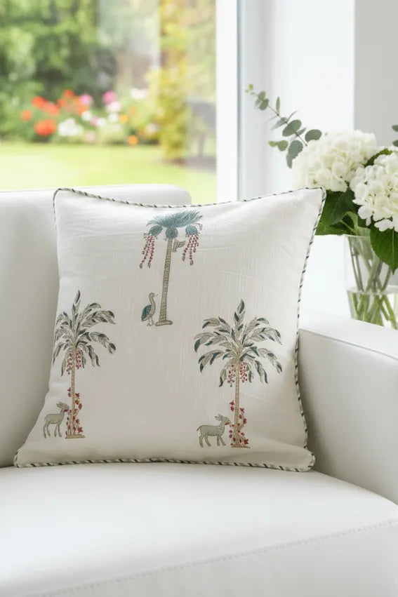 Decorative cushion cover with palm tree design on a white couch with a garden view.