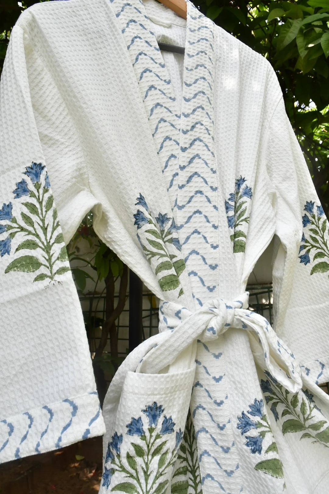 A white waffle-knit robe with blue and green floral and wave patterns, tied at the waist and hanging on a wooden hanger against a background of green foliage.