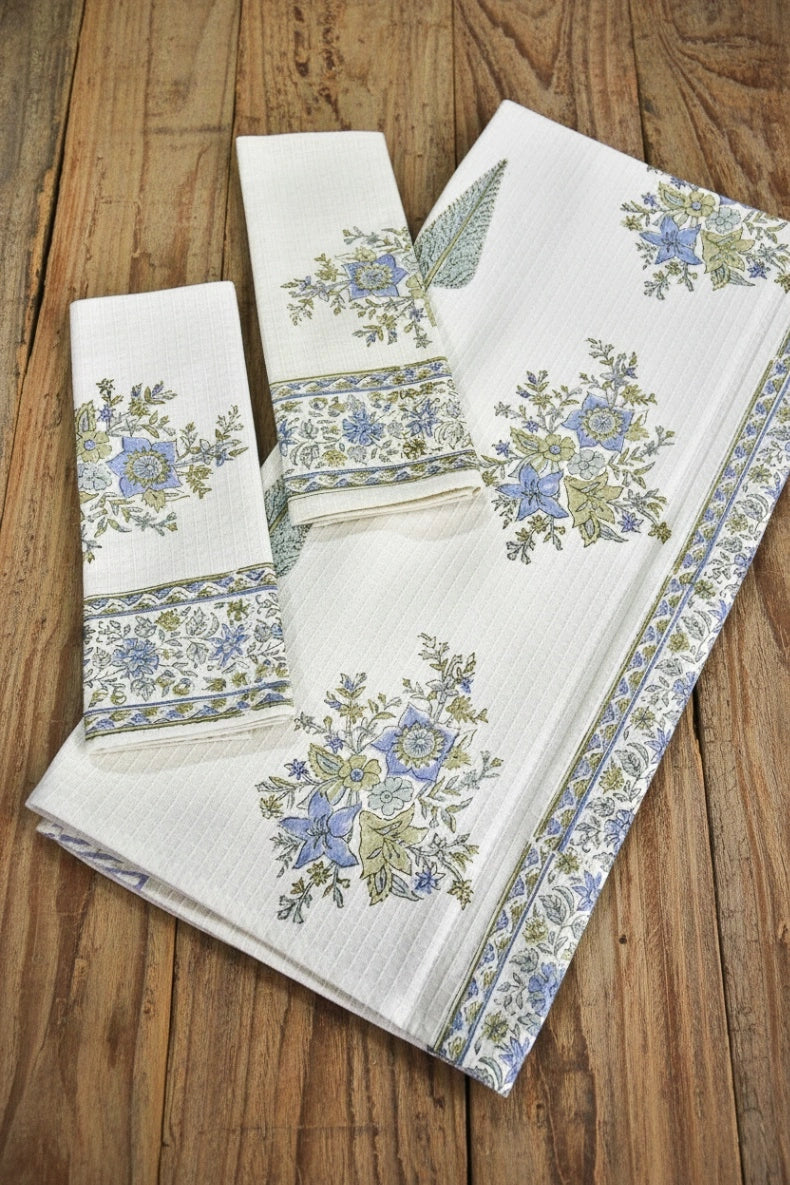 A folded white cotton towel set with blue and green floral prints and border patterns, displayed on a rustic wooden surface.