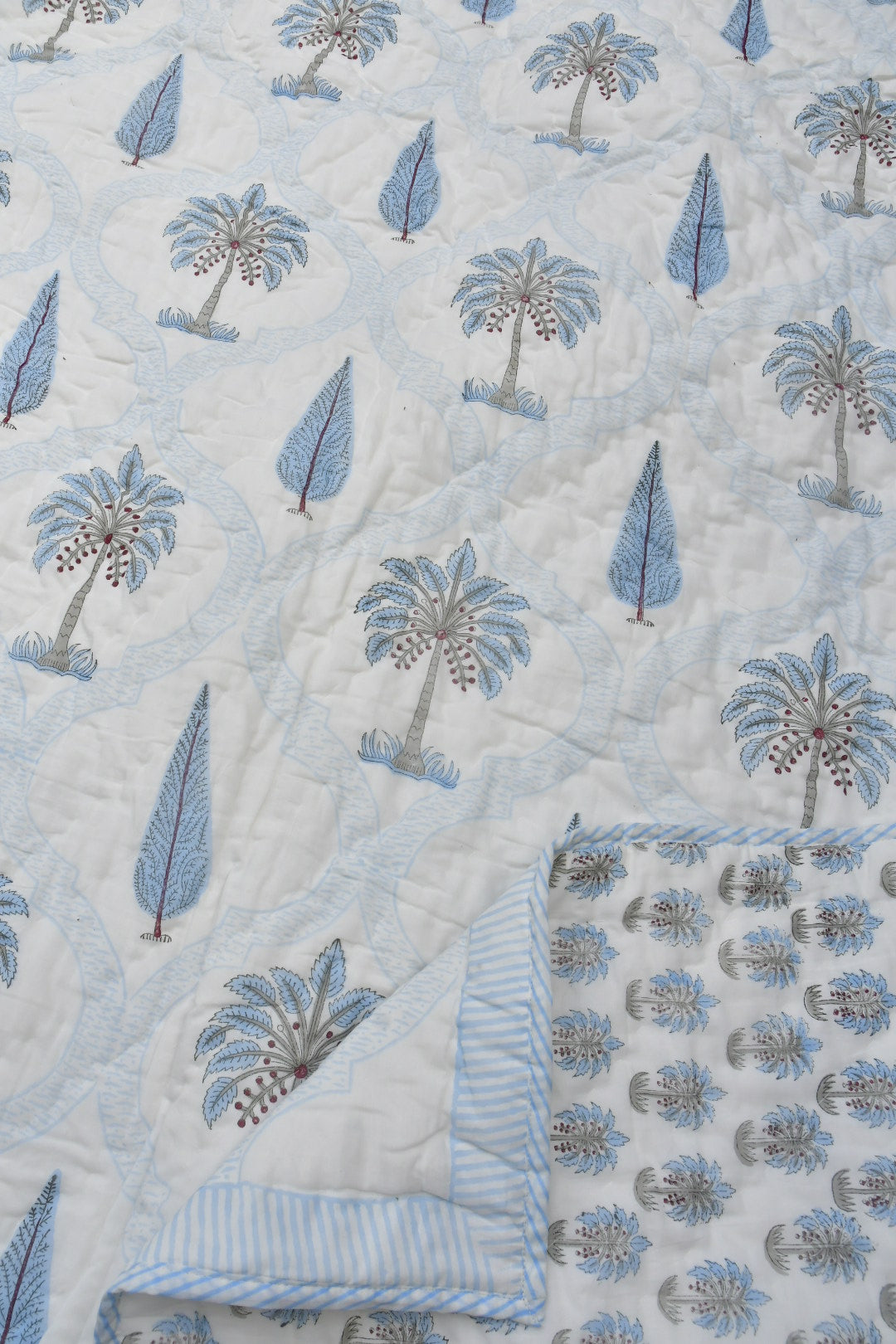 A close-up of a quilted rajai featuring a repeating pattern of blue palm trees and leaf motifs on a white background, with a folded corner showing a light blue and white striped border.