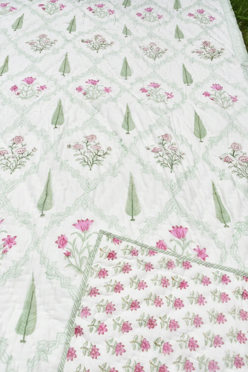 A close-up of a white quilted blanket featuring a repeating pattern of pink flowers and green leaves, with a solid section of dense pink floral embroidery along one corner.