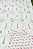 A close-up of a white quilted blanket featuring a repeating pattern of pink flowers and green leaves, with a solid section of dense pink floral embroidery along one corner.