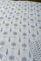 A close-up of a light blue and white printed quilt featuring a repeating palm tree and teardrop pattern, with a folded matching fabric piece placed in the upper right corner.