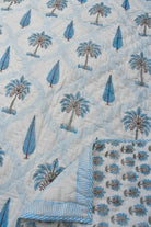 A close-up of a quilted blanket featuring a light blue and white tropical pattern with palm trees and leaf motifs, showing both the front design and the striped reverse side.