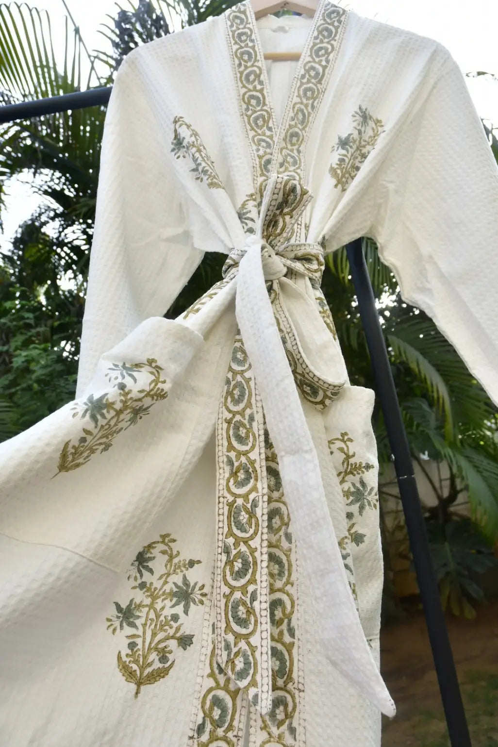 White embroidered garment with gold patterns on a hanger against a blurred green outdoor background