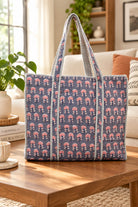 A large woven tote bag with a navy blue base, pink floral block print, and cream striped handles, displayed on a wooden coffee table in a cozy living room setting.