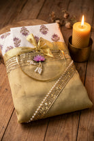 A folded light gold saree with a white and purple patterned border, wrapped with a shiny gold ribbon and bow, and decorated with a small pink lotus flower and white beads.
