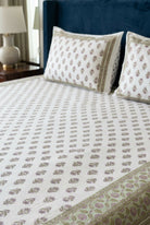 A close-up of a bed made with a white cotton bed sheet and matching pillowcases featuring a repeating brown and green floral block print, set against a dark blue headboard.