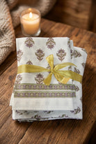 A folded set of white cotton napkins or table linens with a brown and purple floral block print, tied with a shiny gold ribbon on a dark wooden table.
