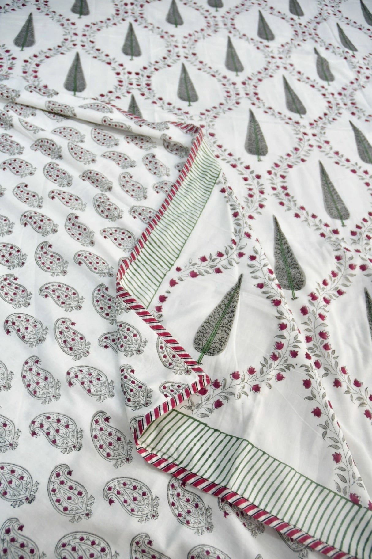 A close-up of a white dohar textile with printed paisley motifs, green teardrop shapes, and red dotted accents, plus a striped border.